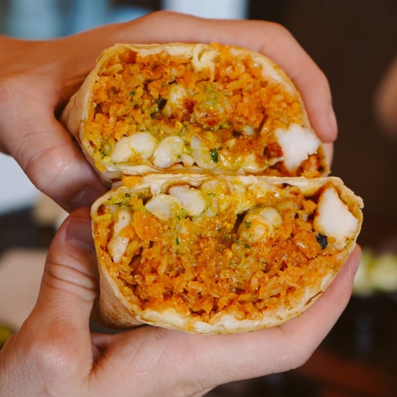 Hands holding a Flying Burro burrito, cut in half, revealing rice, beans, and white cheese.