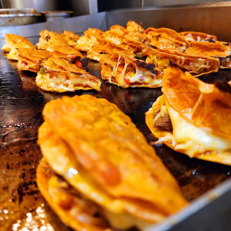 Many cheesy birria tacos cooking on a flat top grill at Flying Burro.