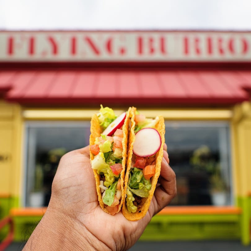 Hand holding two tacos with lettuce, tomato, and radish in front of the Flying Burro restaurant.