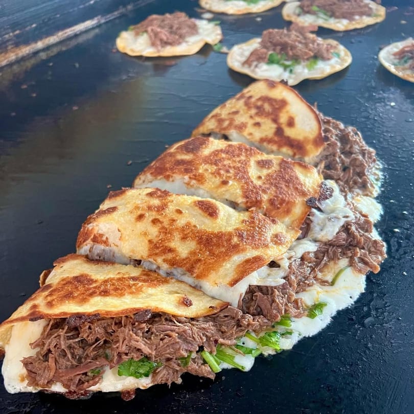 Several Flying Burro quesadillas with shredded beef, cheese, and cilantro cooking on a griddle.