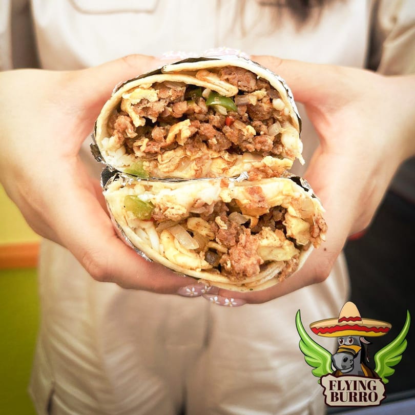 Hands holding a Flying Burro burrito, cut in half, revealing a hearty filling of meat and vegetables.