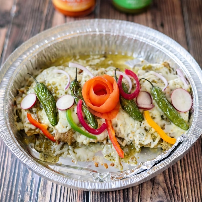Green enchiladas with melted cheese, peppers, onions, and a carrot rose in a foil container.