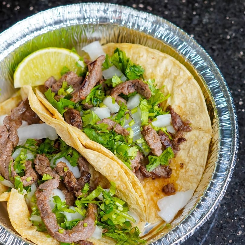 Two carne asada tacos with cilantro, onions, and a lime wedge in a foil container.
