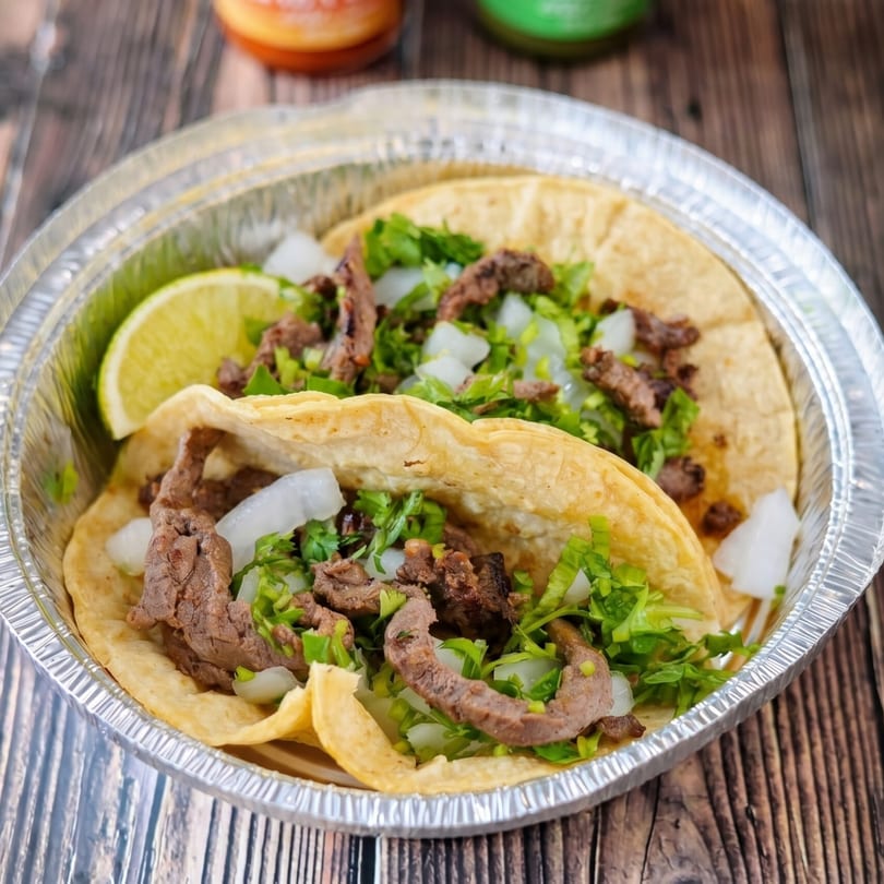 Two carne asada street tacos with onions, cilantro, and lime in a foil container.