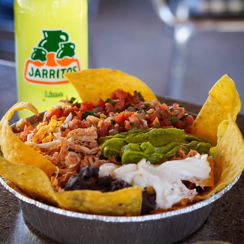 A foil bowl of nachos with chicken, salsa, guacamole, sour cream, and black beans, with a Jarritos soda.