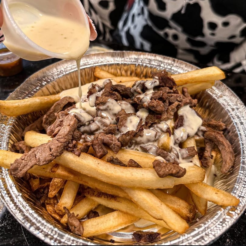 Creamy white sauce being poured over a foil container of golden french fries and steak pieces.