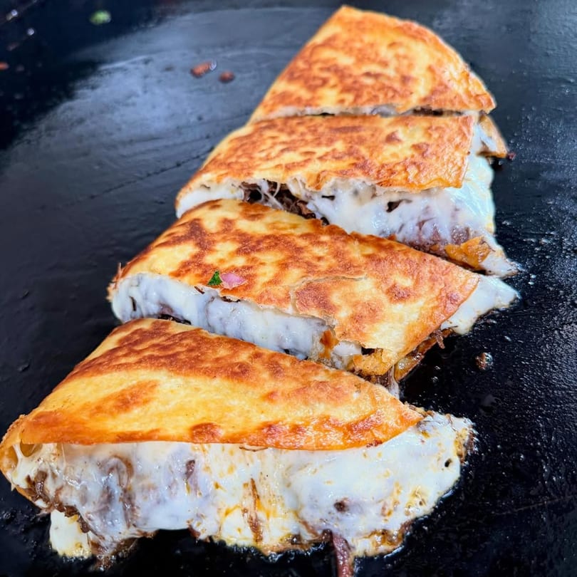 Four cheesy quesadillas with meat filling, cooking on a dark, flat griddle at Flying Burro.