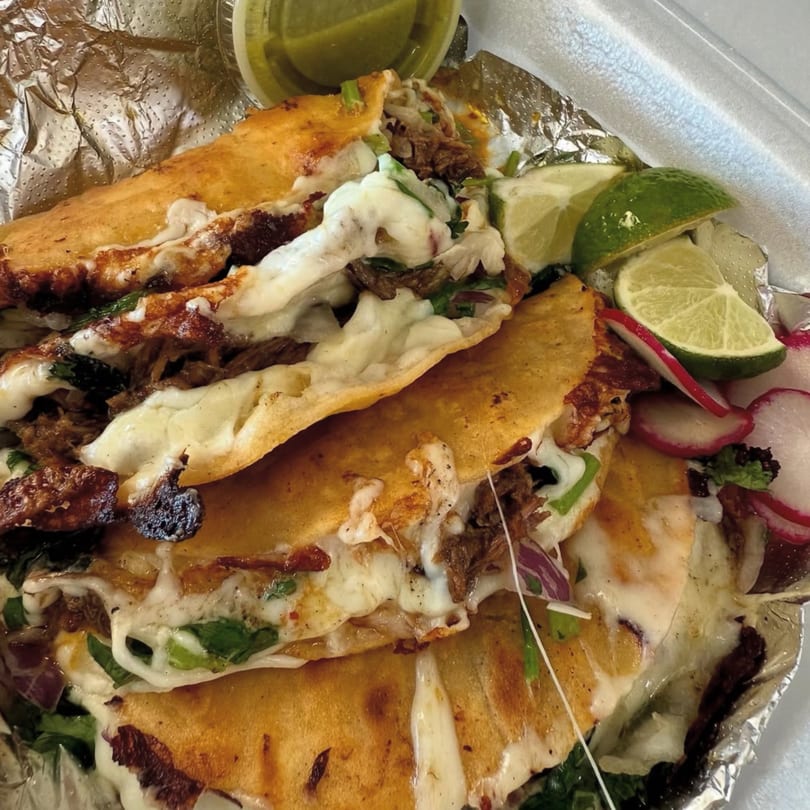 Close-up of three cheesy birria tacos with limes, radishes, and green sauce in a to-go container.