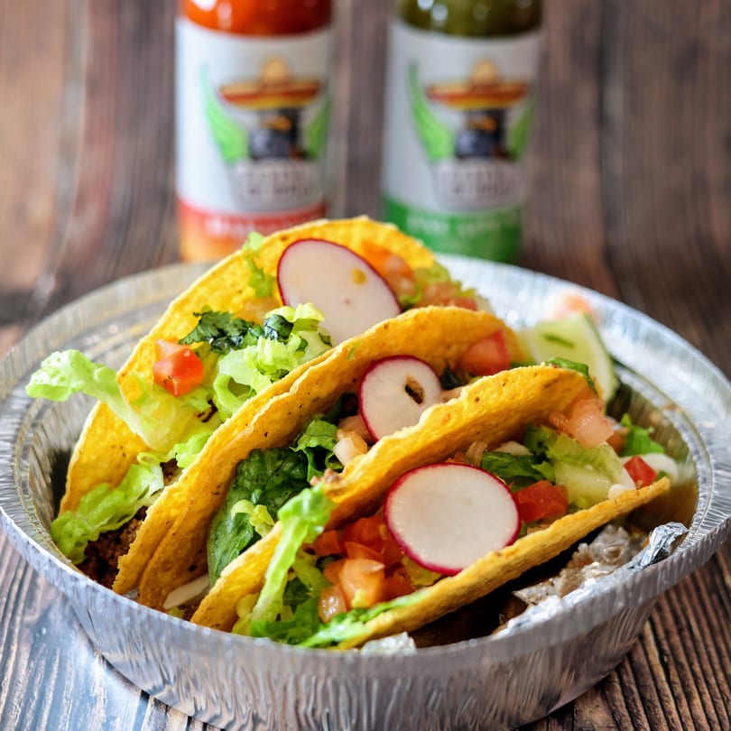 Three Flying Burro tacos with lettuce, tomatoes, and radishes in a foil container.