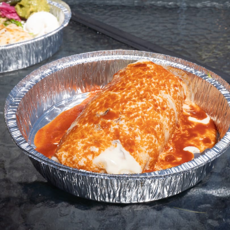 A Flying Burro burrito covered in red sauce and sour cream in a foil takeout container.